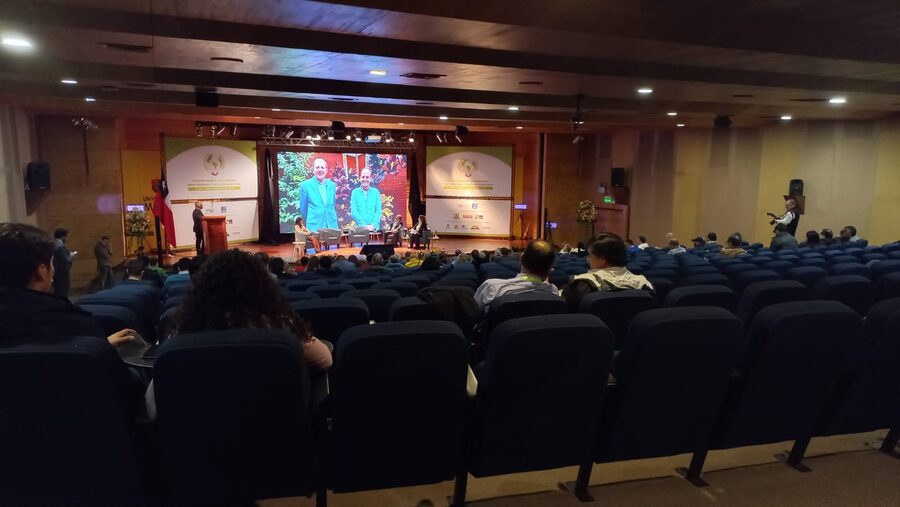 Auditorio con público sentado frente al escenario y pantalla con la transmisión del panel