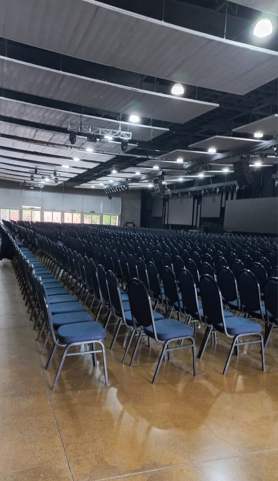 Salón de congresos vacío con cientos de sillas alineadas y sistema de audio line array suspendido del techo