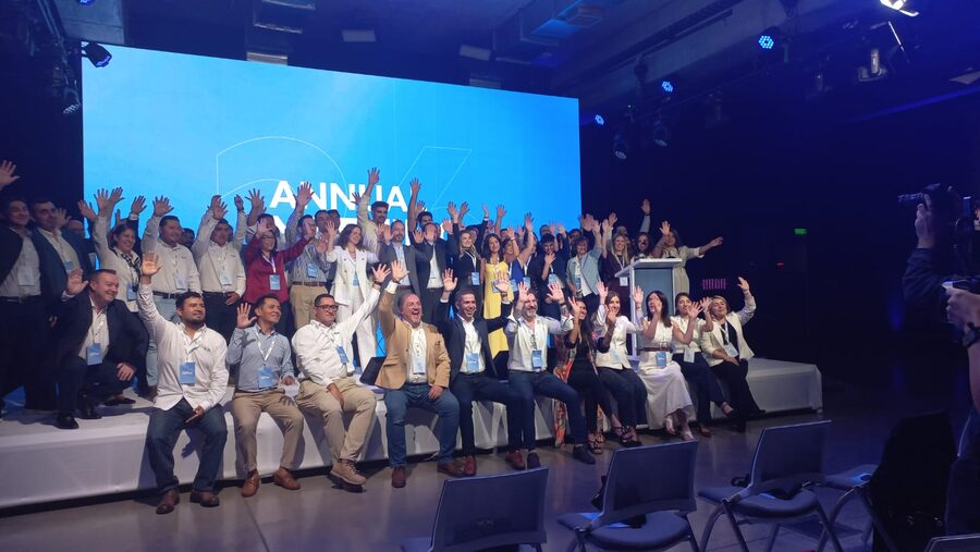 Fotografía grupal del equipo sobre el escenario con los brazos en alto, frente a la pantalla LED de la convención